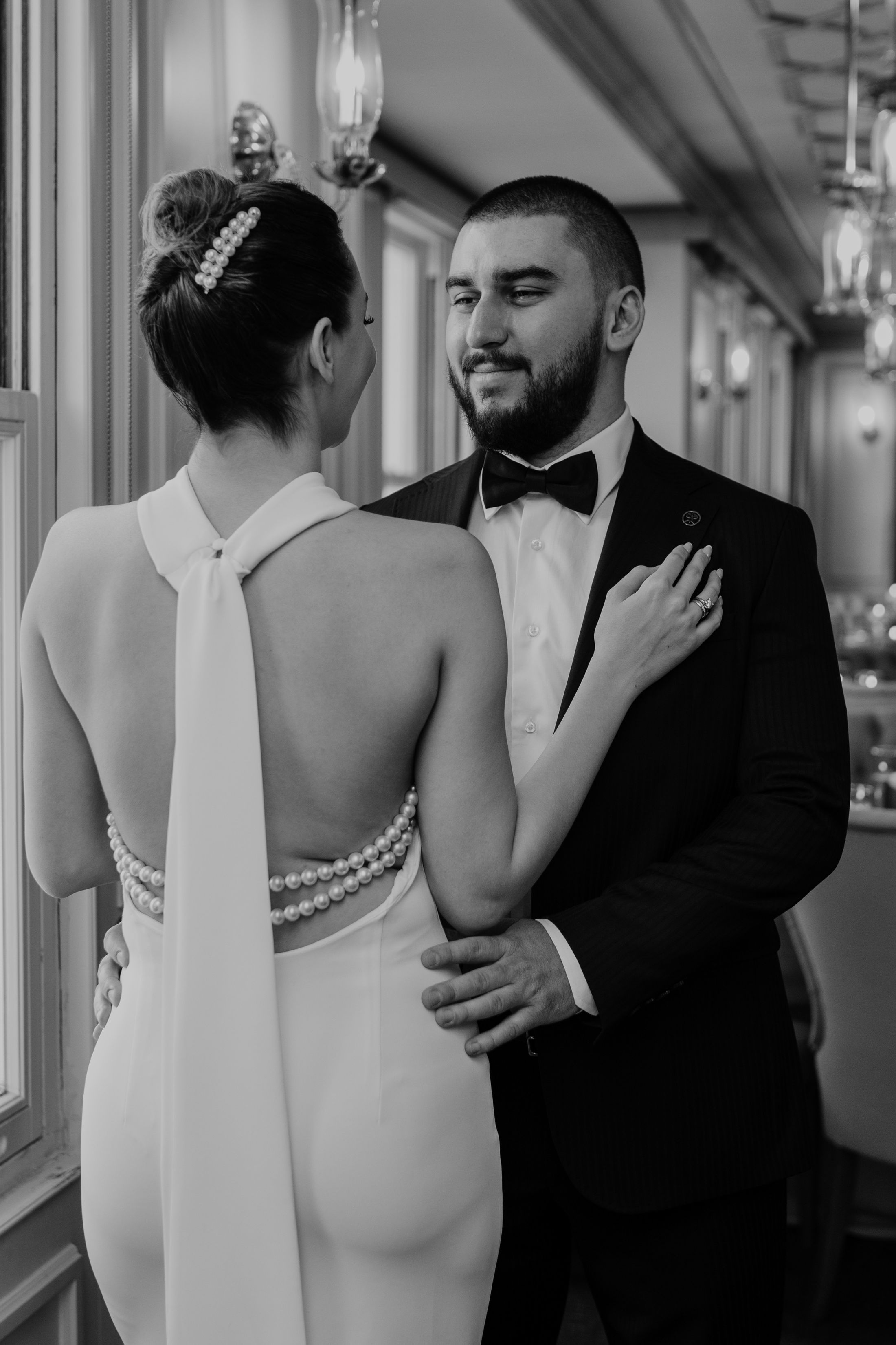 Sepasang pengantin berdekapan dalam foto hitam putih; pengantin laki-laki berdasi kupu-kupu tersenyum, pengantin perempuan bergaun punggung terbuka