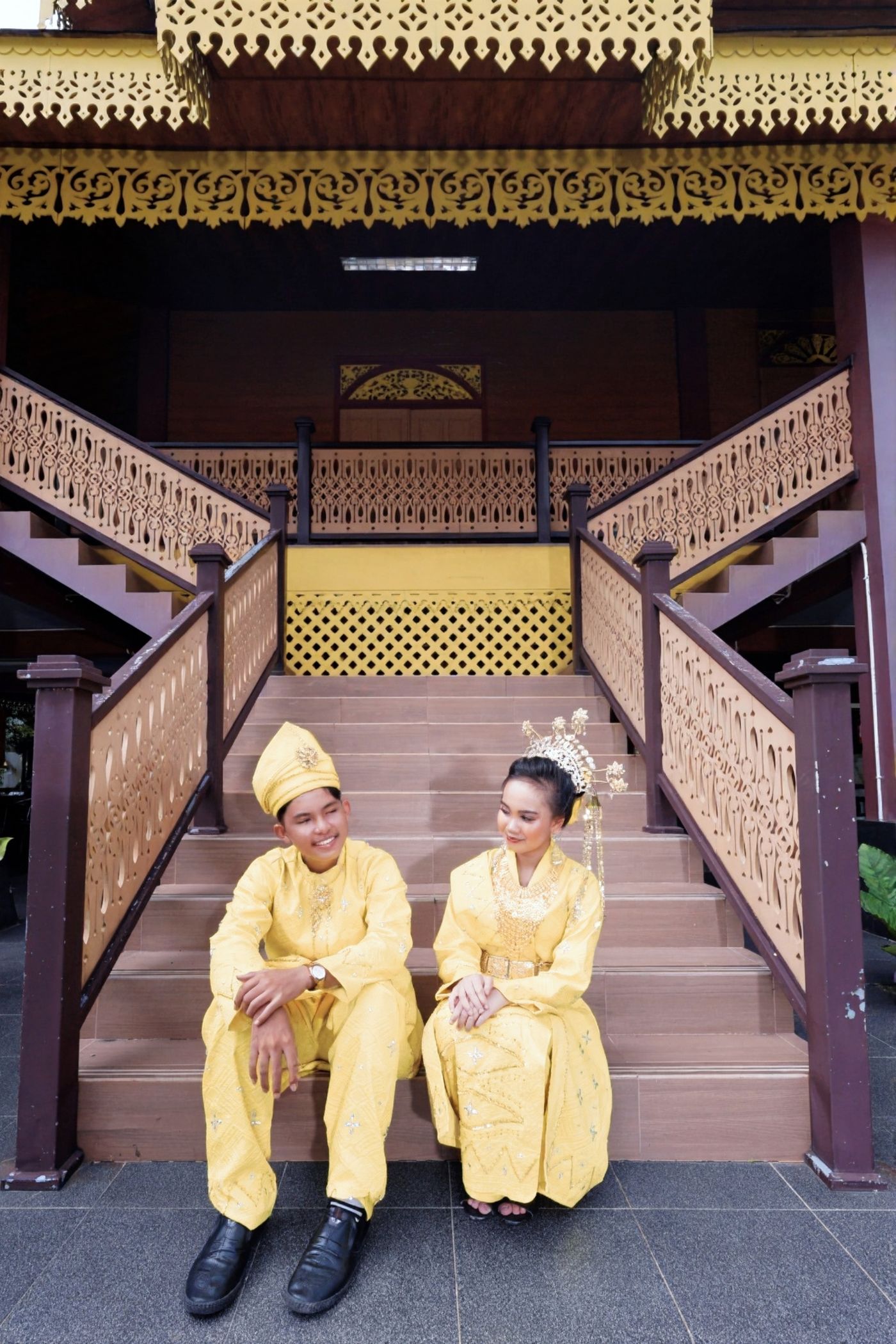 Pasangan berbusana kuning di tangga kayu dengan ukiran Melayu